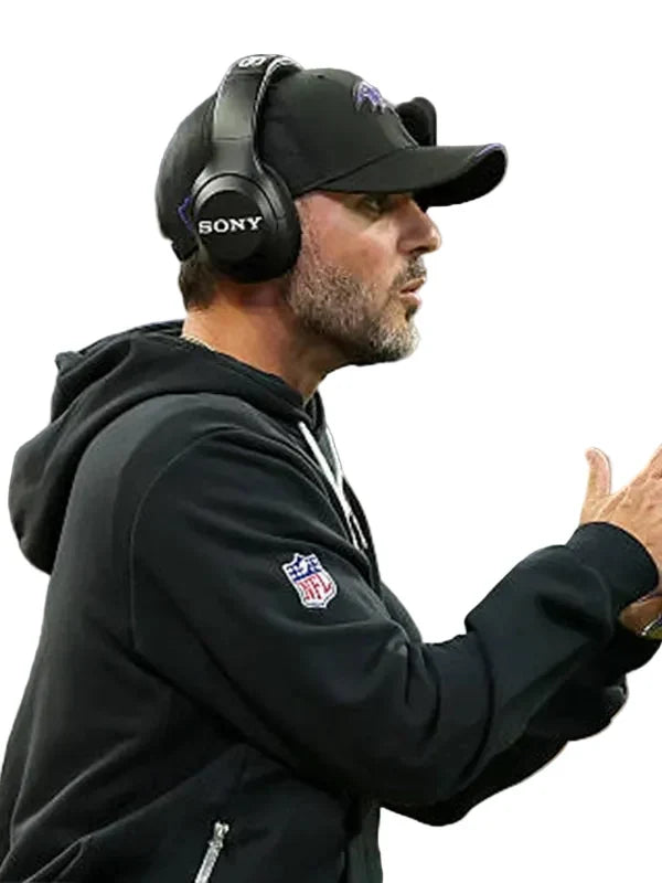A man in a black NFL hoodie and black cap wearing a black Sony over-ear headset, shown in profile against a blurred stadium background.