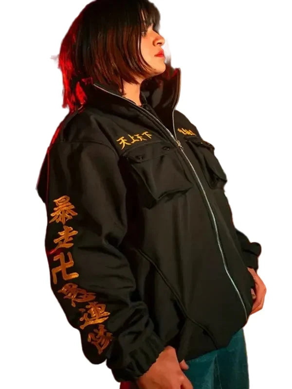 A person standing outdoors at night wearing a black zip-up jacket with large gold Chinese characters printed down one sleeve and across the chest, paired with blue jeans.