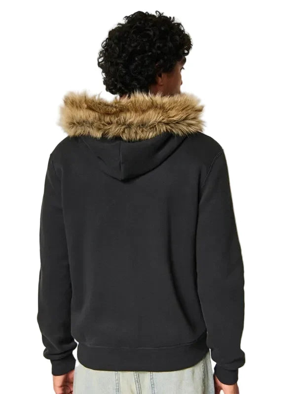 A person shown from the back wearing a black pullover hoodie with a tan faux-fur trimmed hood, against a plain light beige studio background.