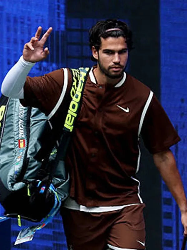 A male tennis player wearing a brown Nike athletic set with white trim, carrying a large Babolat racket bag over his shoulder, set against a dark blue background.