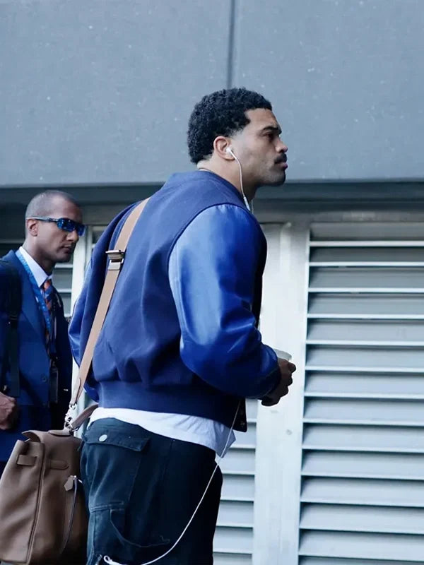 A man wearing a blue leather-look bomber jacket, dark cargo pants, and a brown shoulder bag walks outdoors next to a gray building with louvered windows. Another man in sunglasses is partially visible in the background.