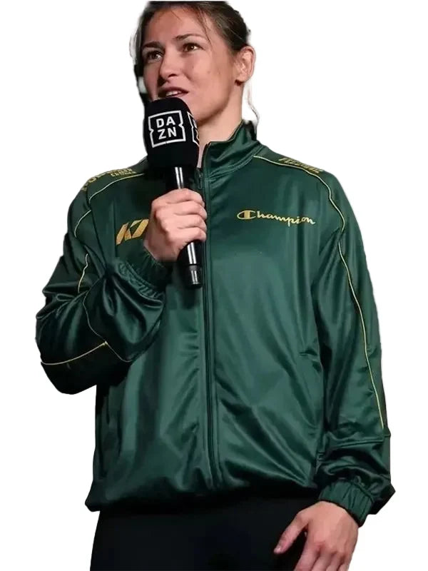 A woman wearing a dark green Champion track jacket with gold piping and logo, holding a black microphone with a DAZN logo, against a solid light gray background.