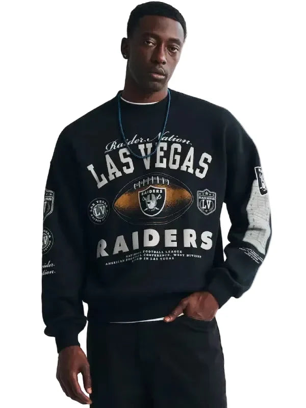 A man wearing a black Raiders Nation Las Vegas Raiders crewneck sweatshirt with white and gold graphics, paired with black pants, against a light gray studio background.
