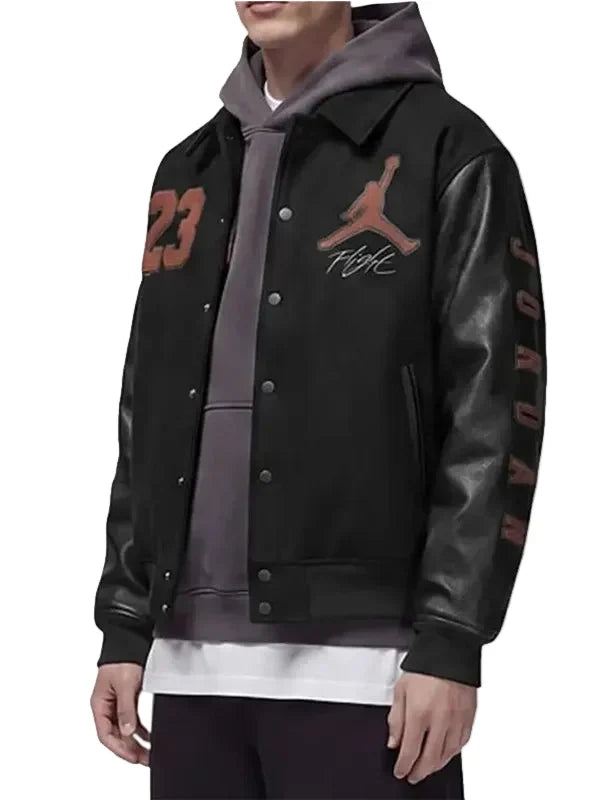A person wearing a black varsity-style jacket with leather-look sleeves, a gray hoodie underneath, and brown Jordan number and logo graphics on the chest and sleeve.