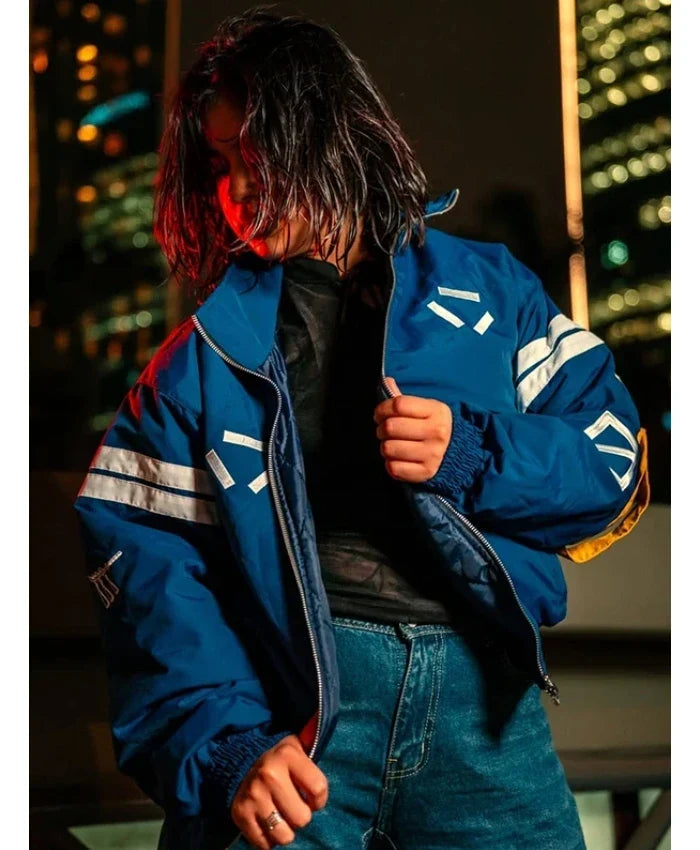 A person wearing a blue bomber jacket with white graphic stripes and a black shirt, posed against a blurred nighttime cityscape background.