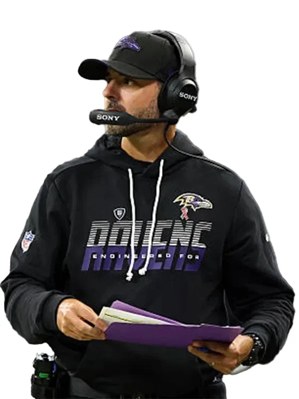 A man wearing a black Baltimore Ravens hoodie with 'DAIENCE ENGINEERED FOR' text, a black cap, and a Sony headset, holding purple folders against a light gray background.