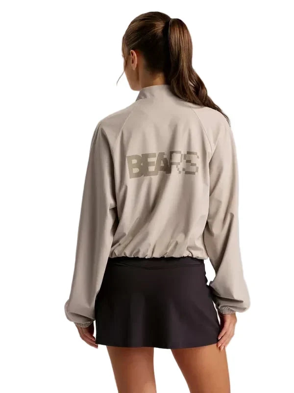 A woman shown from the back wearing a light beige long-sleeve athletic jacket with a large dark gray 'BEARE' logo on the back, paired with a dark skirt, against a solid light gray background.