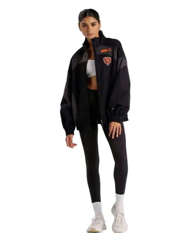 A woman modeling a black athletic jacket with gray side panels and orange logo details, paired with black leggings, white socks, and white sneakers, against a solid light gray background.