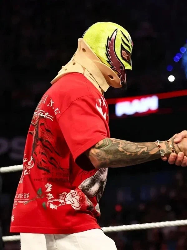A wrestler in a bright neon yellow mask with black and red details, a red graphic t-shirt, and white pants shakes hands inside a wrestling ring under arena lights.