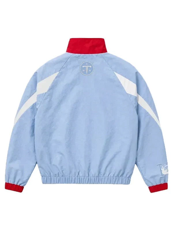 A light blue windbreaker jacket shown from the back, featuring a red collar and cuffs, white diagonal shoulder stripes, and a circular 'TT' logo on the upper back.