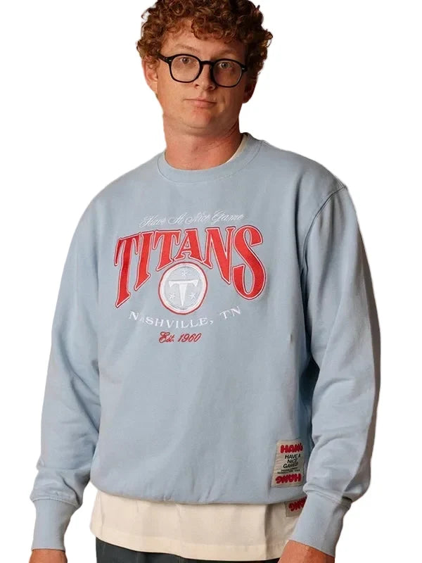 A man wearing a light blue sweatshirt with a red and white 'Titans' graphic on the front, paired with a cream undershirt and black glasses, against a light gray background.
