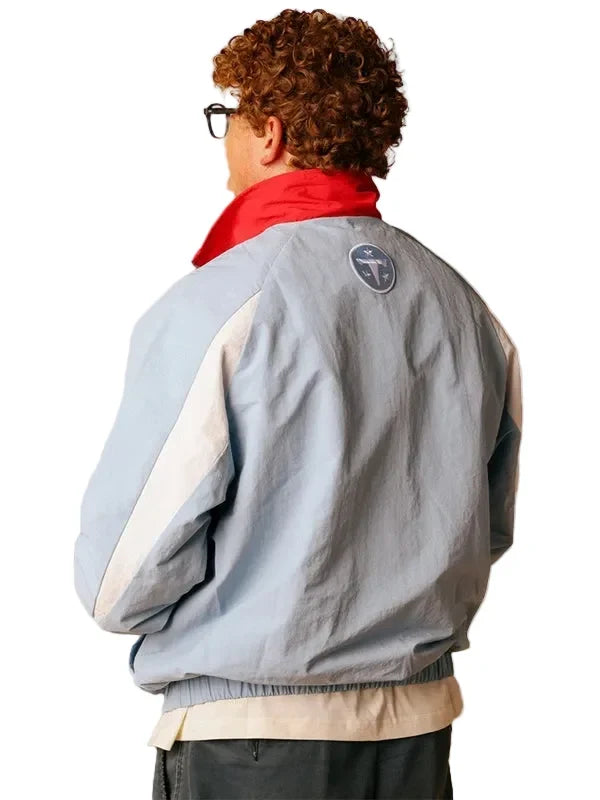 A person shown from the back wearing a light blue color-blocked jacket with cream and red panels, a red neck collar, and a circular logo patch on the upper back.